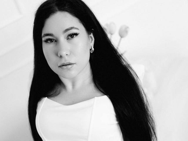 Zwart-wit foto van AnnabellDeltoro, met lang donker haar, die een witte top draagt en recht in de camera kijkt. met een neutrale uitdrukking. Hoog contrast, scherpe focus op het gezicht, zachte achtergrond.