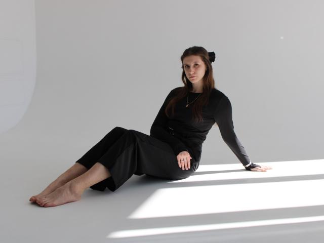 LilianaShine sits on the floor with legs extended, leaning back on her right hand. She wears a black long-sleeve dress and has her hair in a bun. Bright light casts shadows. Her feet are bare.