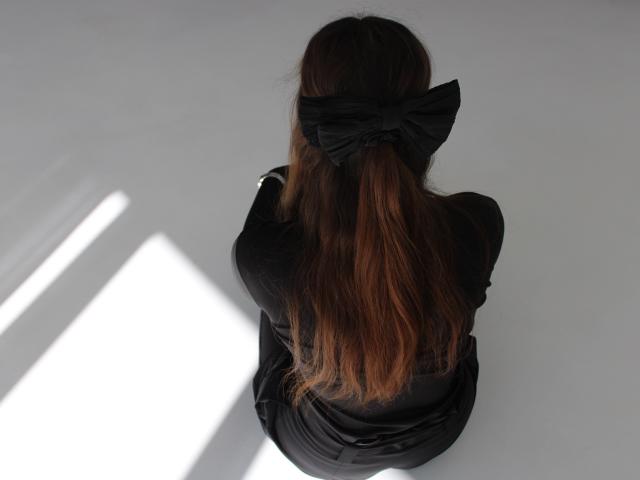 LilianaShine with long brown hair tied in a black bow, sitting on the floor, back to the camera, wearing a black dress. Bright white light casts sharp shadows on her.