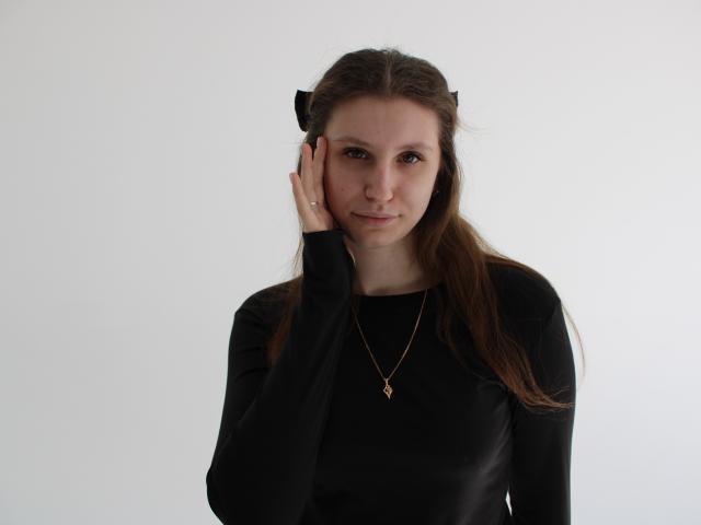 Fotografia di LilianaShine con capelli castano scuro, che indossa una maglia nera a maniche lunghe e una collana dorata. Lei tocca il viso con la mano destra, sfondo bianco, tensione sessuale, posa casuale..