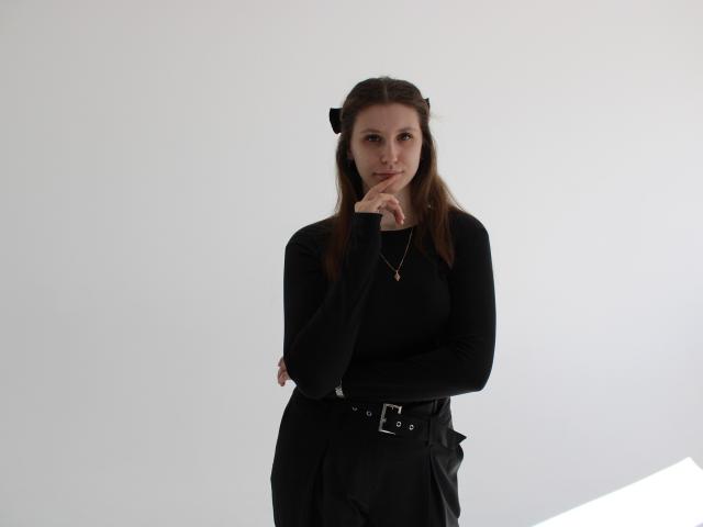 Photographie de LilianaShine avec de longs cheveux bruns, portant un haut noir à manches longues et un pantalon noir, debout devant un fond blanc. Elle se mord la doigt avec une expression sérieuse. Putain, elle est sexy..