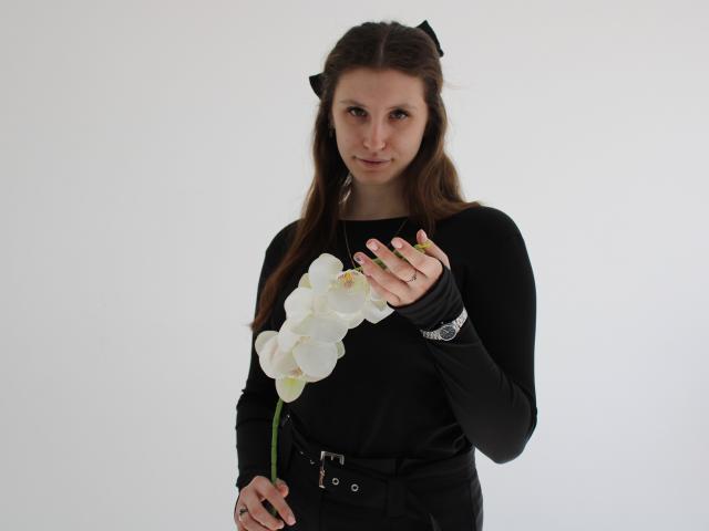 Fotografía de LilianaShine sosteniendo una orquídea blanca con ambas manos, vistiendo una blusa negra de manga larga, con cabello oscuro. Diadema con lazo negro y cinturón negro. Fondo blanco. Expresión muy seria..