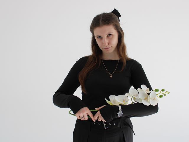 Photographie de LilianaShine avec de longs cheveux bruns, un haut noir à manches longues, une jupe noire, tenant des lys blancs, un nœud noir dans les cheveux. cheveux, collier en argent, léger sourire, fond blanc, ombres minimales, tension sexuelle.