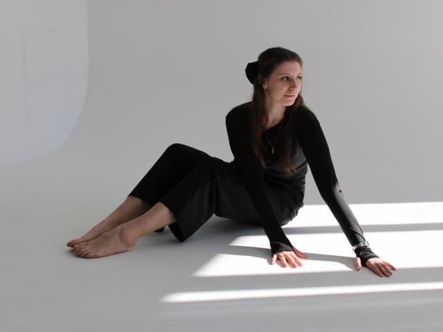 LilianaShine sits on the floor, legs extended, leaning on her right arm. She's wearing a black long-sleeve shirt and pants, barefoot, with shadows on the white floor. She's looking to the side.