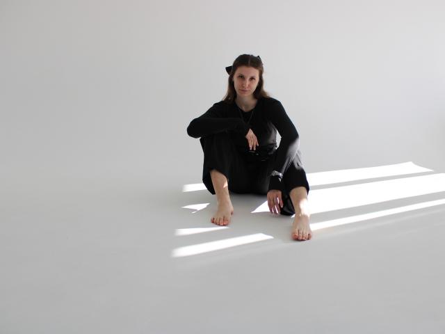 LilianaShine está sentada en el suelo con las piernas separadas, vestida con un conjunto negro. Está descalza, con las manos apoyadas en las rodillas, y está a la sombra de una luz brillante. Su expresión es neutra, ligeramente intensa..