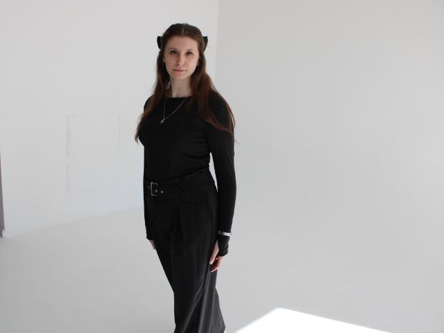 LilianaShine est debout devant un fond blanc, vêtue d'un haut noir à manches longues et d'un pantalon noir avec une ceinture. Elle a de longs cheveux bruns, une expression sérieuse et un petit accessoire pour les cheveux noir..