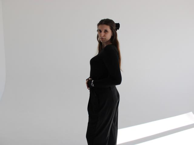 LilianaShine steht seitlich in einem schwarzen Kleid, ihr langes, braunes Haar ist zu einem Dutt gebunden, vor einem schlicht weißen Hintergrund. diagonales Lichtstreifen. Sie blickt über ihre Schulter und lächelt..