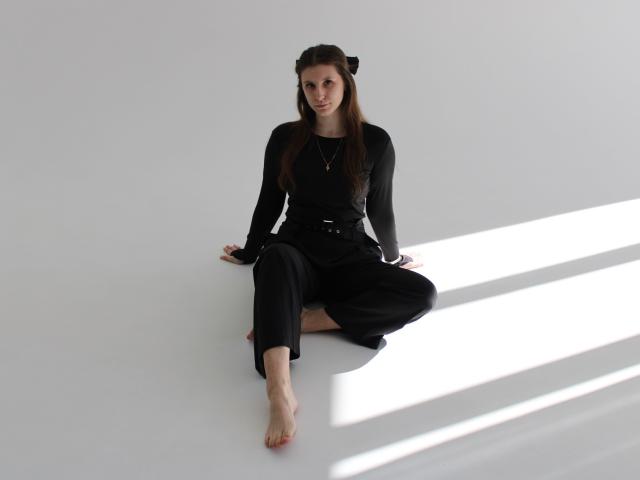 LilianaShine, assise en tailleur sur un sol blanc, portant un haut noir à manches longues et un pantalon, pieds nus, avec de longs des cheveux bruns et une pince à cheveux noire, des ombres projetées sur sa droite.