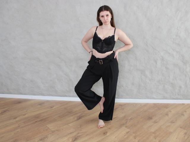 LilianaShine está em pé com confiança, com uma perna dobrada, as mãos na cintura, vestindo uma blusa preta com detalhes em renda e calças de pernas largas. Descalço no chão de madeira.
ela exala tensão sexual, olhando diretamente para a câmera..