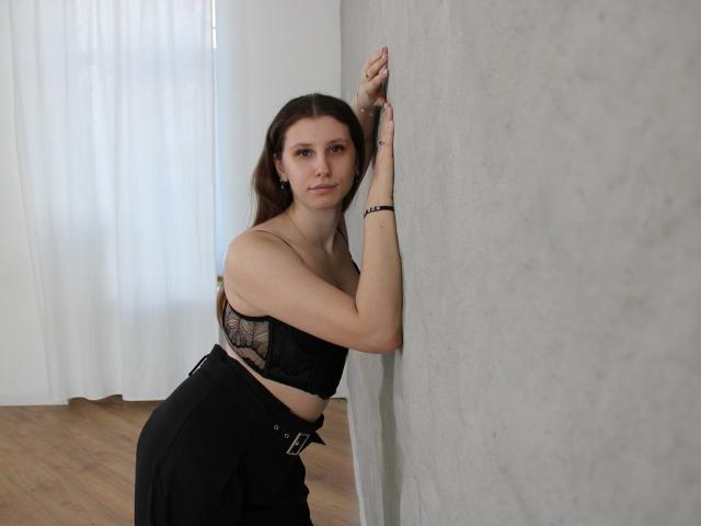 LilianaShine, com cabelo escuro e pele clara, está encostada em uma parede branca, vestindo um sutiã de renda preta e uma saia de cintura alta. Criando uma pose provocante com uma expressão sedutora. Luz brilhante que entra pela janela.