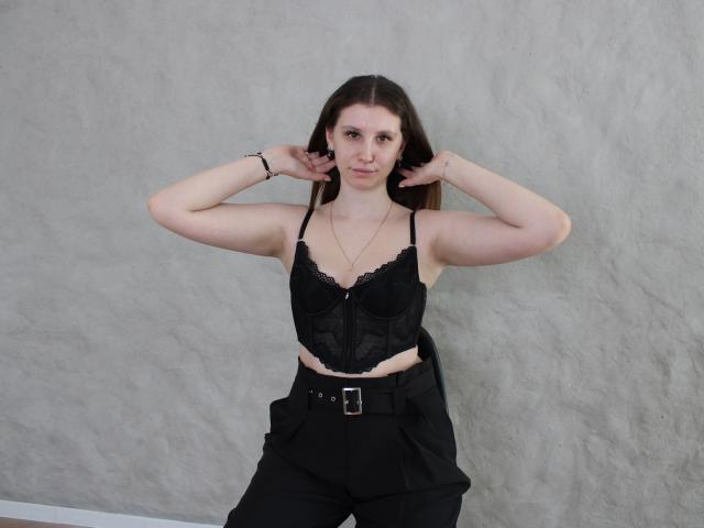 LilianaShine, de pele clara e cabelos castanho-escuros, está em frente a uma parede cinza com textura. Ela veste uma blusa preta de renda, calças pretas de cintura alta com um cinto e um colar. Braços levantados, mãos atrás da cabeça. Incrivelmente sexy..