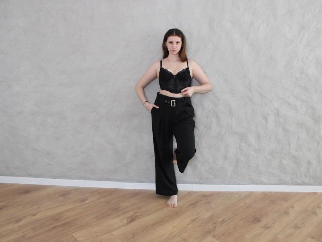 LilianaShine è in piedi a piedi nudi contro un muro grigio con una superficie ruvida, indossando una camicetta di pizzo nero e pantaloni neri larghi. cintura.
lei si appoggia in modo rilassato, con una mano appoggiata al fianco. Pavimento in legno..