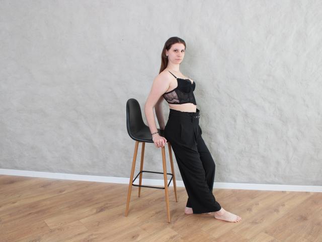 LilianaShine, met een bleke huid en donkerbruin haar, leunt tegen een zwarte stoel, gekleed in een zwarte kanten beha en een loszittende zwarte broek aan, blote voeten op houten vloer, grijze, gestructureerde muur, duidelijke seksuele spanning..