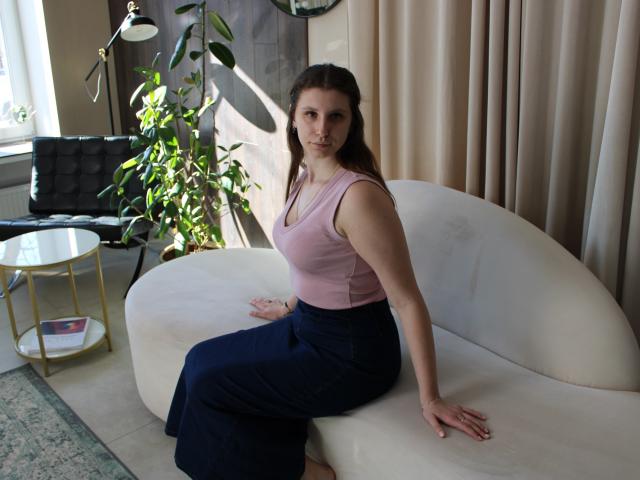 LilianaShine, une femme aux cheveux foncés, avec une poitrine de taille moyenne, portant un débardeur rose et un pantalon noir, assise sur un canapé blanc incurvé, baignée de soleil. salon, plante en arrière-plan, ombre sur le mur, pose décontractée, ambiance séduisante..