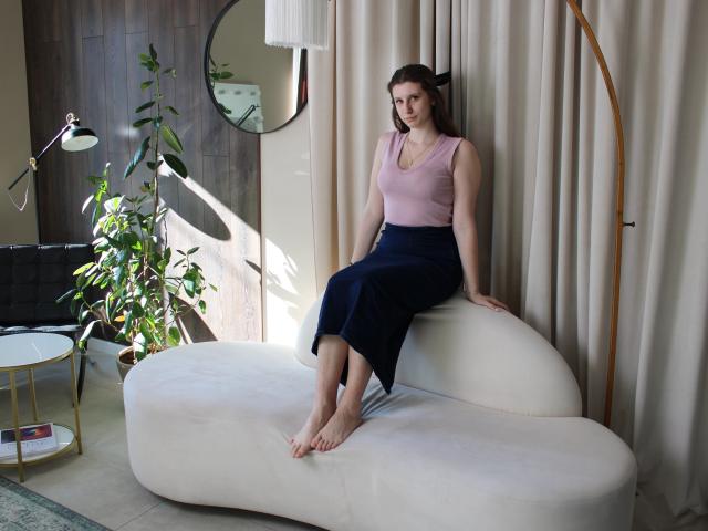 LilianaShine sits on a white, curvy couch, legs crossed, wearing a pink tank top and black skirt. Sunlight streams in, casting shadows. Plants and a modern lamp in the background.