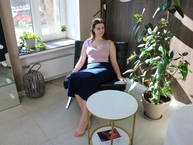 LilianaShine è seduta su una sedia nera, indossa una canotta rosa e una gonna nera, con le gambe divaricate e i piedi nudi, vicino a un tavolo bianco. e una pianta in vaso. La luce solare intensa la illumina..