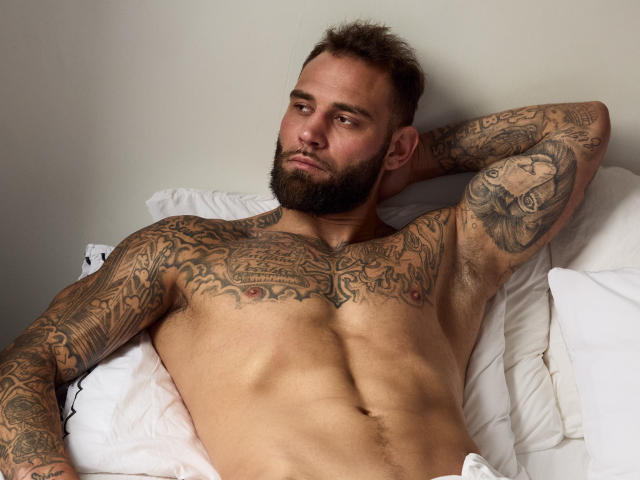 Photograph of a heavily tattooed AlphamanJay with a muscular build, lying on white pillows, arms behind head, chest and arms covered in detailed black ink, bearded, intense gaze, casual pose.