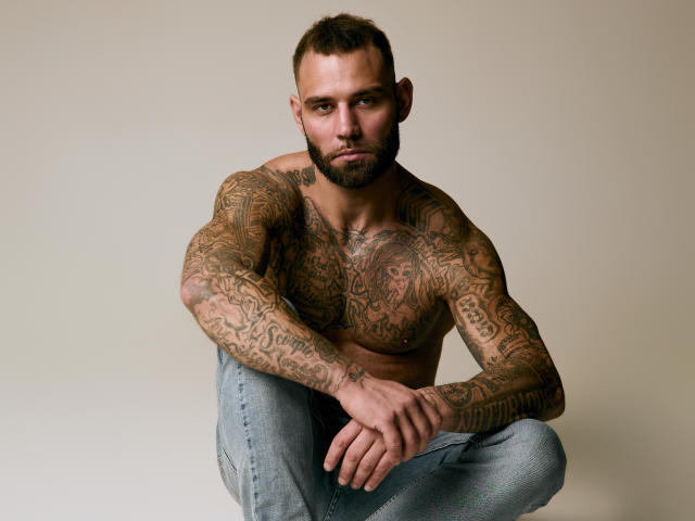 Fotografia de AlphamanJay sem camisa, com extensas tatuagens cobrindo seu peito e braços musculosos, agachado com as pernas. cruzado, vestindo calças jeans azuis, cabelo curto e escuro, olhar intenso, barba rústica..