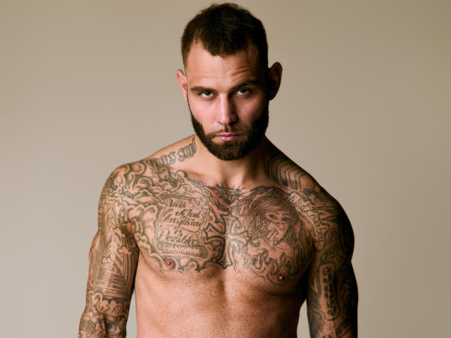 Fotografie eines muskulösen, bärtigen "Alphamanns" namens Jay, mit ausgedehnten Tattoos, die seine Brust und Arme bedecken. Er ist oben ohne, blickt intensiv mit ernstem Gesicht und hat eine deutlich sichtbare, erigierte Brustwarze..