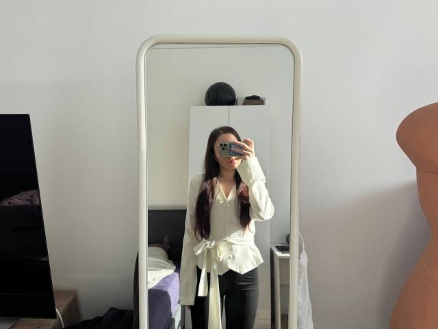 Zelfportret van AnnabellDeltoro met lang donker haar, gekleed in een witte blouse en zwarte broek, staand voor een spiegel, met een telefoon in haar hand, zonlicht verlicht haar. Spiegelbeeld, slaapkamer setting.