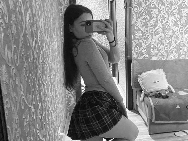 Fotografía en blanco y negro de AnnabellDeltoro arrodillada, frente a un espejo, tomando una selfie. Ella lleva una blusa ajustada y una falda escocesa, que resaltan sus glúteos. Fondo: cortinas con estampado, sofá con un bolso. Ligera tensión sexual..