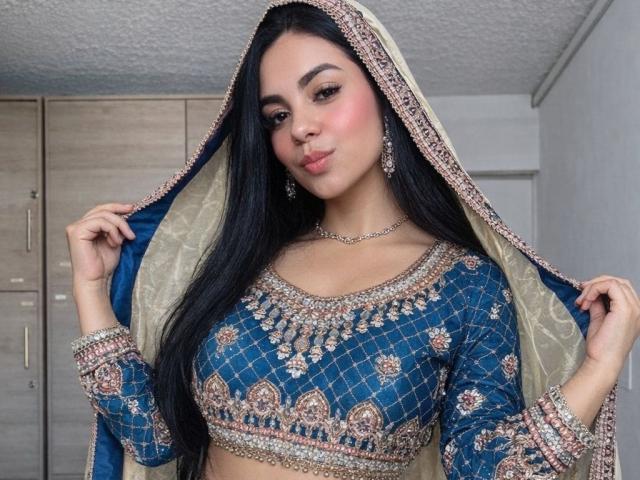 DanMartines, com cabelos longos e pretos, vestindo uma blusa azul bordada e um véu, segurando o véu, olhando para a câmera. com os lábios levemente curvados para dentro. Joias brilhantes, pose sexy e confiante..