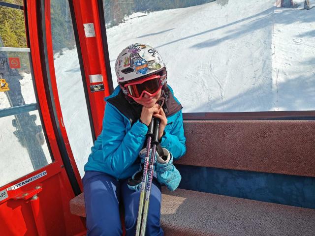 SofiaLiub sits on a red ski lift seat, wearing a blue jacket, black pants, and a white helmet with red goggles. Holding ski poles, gazing into the snowy landscape.