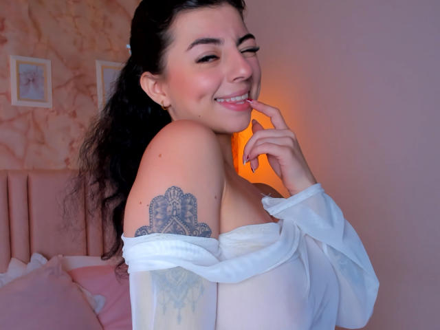 Anntonella Watson, con i capelli scuri, sorride in modo seducente, mordendosi il dito. Indossa una maglietta bianca a spalle scoperte, che rivela un grande tatuaggio a forma di mandala sul braccio. Illuminazione in tonalità rosa e arancione tenui..