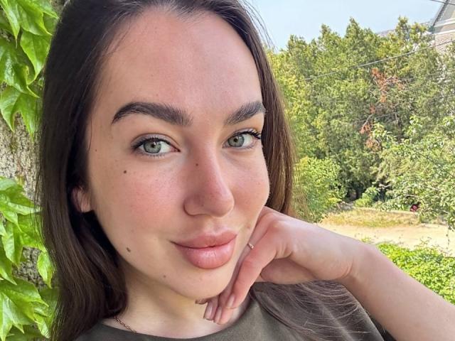 Close-up of AraiaSheri, a fair-skinned woman with green eyes, dark brown hair, and small moles on her face. She's leaning against a tree with green leaves, smirking slightly. Outdoor background.