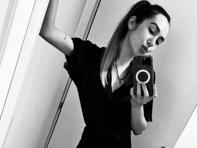 Black-and-white selfie of AraiaSheri, holding a phone with a ring grip, wearing a black top, long hair in a ponytail, moles on her cheek, arm raised, seductive gaze.