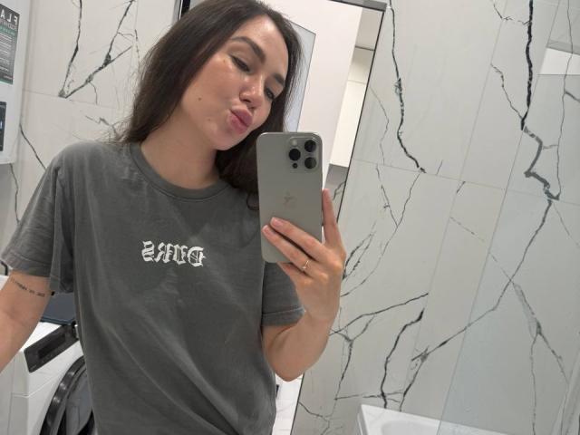 AraiaSheri, com seus longos cabelos escuros, faz beicinho para o espelho enquanto segura um iPhone. Ela veste uma camiseta cinza folgada com a palavra "RAT" em letras brancas. Azulejos com padrão de mármore ao fundo.