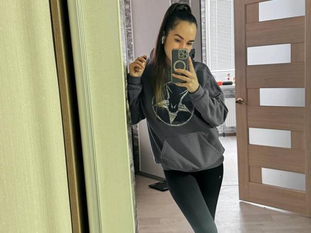 AraiaSheri takes a mirror selfie, holding a phone with a green case. She's wearing a gray hoodie with a graphic, black pants. Her hair is in a high ponytail. Fucking sexy.