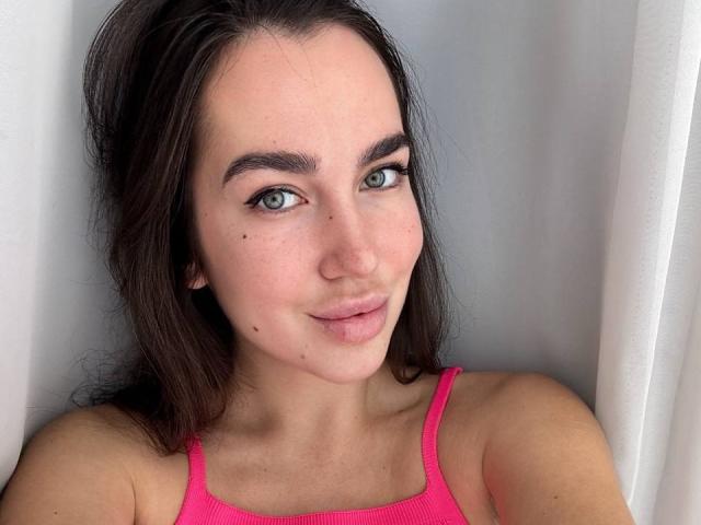 Close-up of AraiaSheri, a fair-skinned woman with dark brown hair, blue eyes, and small moles on her cheeks. She's wearing a bright pink tank top, looking directly at the camera with a slight smirk.