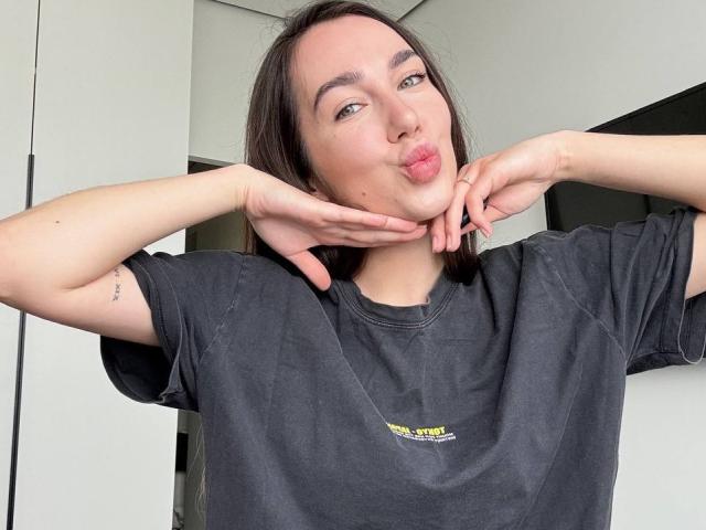 AraiaSheri with long black hair, puckering lips, and hands framing face. Wearing a black T-shirt with small yellow graphic. Pale skin, visible tattoo on left forearm. Sexual tension from provocative pose.