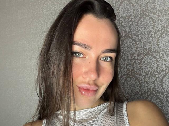 Close-up of AraiaSheri, a young woman with long dark brown hair, blue eyes, and slightly parted lips showing a hint of saliva. She's wearing a white mesh top against a patterned background.