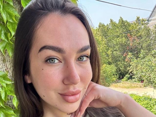Close-up of AraiaSheri, a young woman with long dark hair, green eyes, and light skin, resting her chin on her hand, outdoors with lush greenery in the background. She's looking directly at the camera with a slight smile.