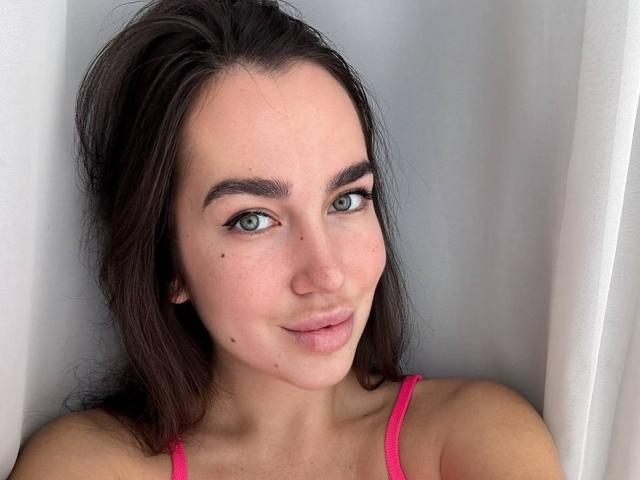 Close-up of AraiaSheri, a young woman with long dark brown hair, blue eyes, and a subtle smile. She's wearing a pink spaghetti-strap top, looking directly at the camera with a sexy gaze.