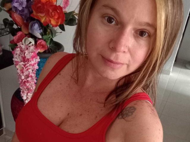 Close-up of PamelaColin, a fair-skinned woman with brown hair, wearing a red tank top, showing cleavage, with a floral arrangement in the background. She's looking at the camera with a subtle smile.