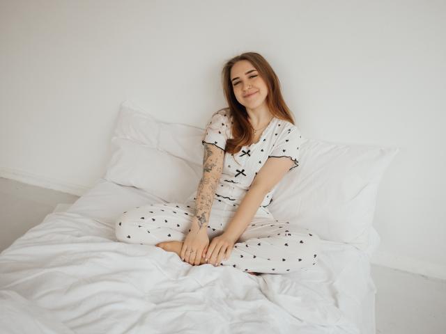 LauraHlot, avec ses longs cheveux bruns, est assise sur un lit blanc, vêtue d'une robe blanche à pois, souriante, les jambes croisées, et tatouée. bras visible, fond blanche et lumineuse, décontracté, sexy, pose détendue..