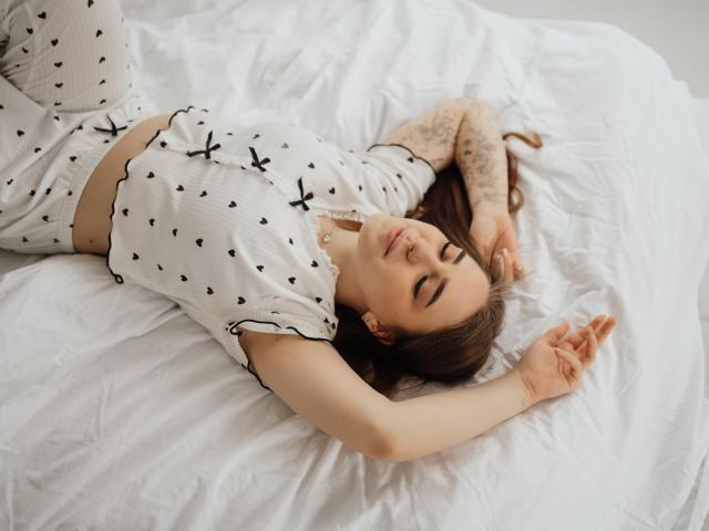 LauraHlot lies on a white bed, arms above her head, wearing a white, black-leaf-patterned top and pants. She has tattoos on her arm, eyes closed, looking relaxed. Bright lighting.