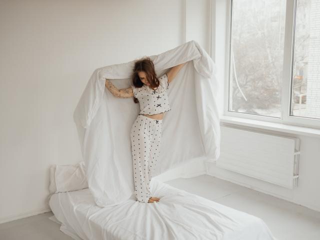 LauraHlot with curly brown hair, wearing white polka-dot pajamas, standing on a white bed, holding a large white duvet, sunlight streaming through a window, casting soft shadows. Fucking sexy.