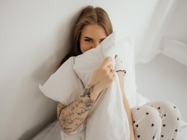 LauraHlot, mit braunen Haaren und einem tätowierten Arm, umklammert ein weißes Kissen und versteckt sich dahinter. Sie trägt eine weiße Hose mit Herzmuster und blickt verführerisch in die Kamera. Helles Licht, weiche Schatten.