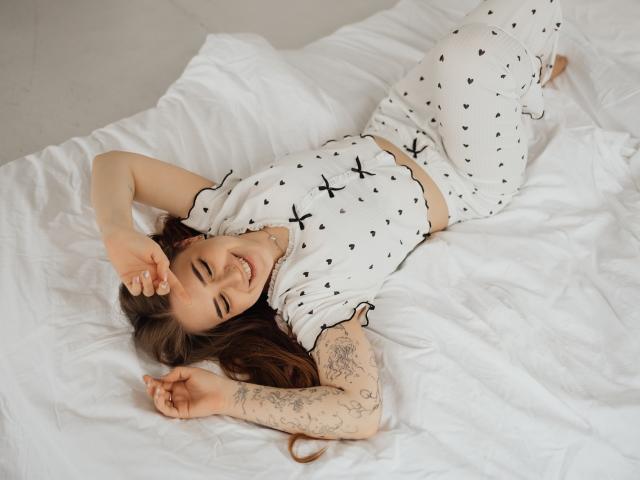 LauraHlot está acostada sobre sábanas blancas, vistiendo un pijama blanco con cruces negras. Ella está sonriendo, con los brazos levantados, mostrando su antebrazo tatuado. Su cabello oscuro contrasta con la tela blanca. Ambiente muy relajado..