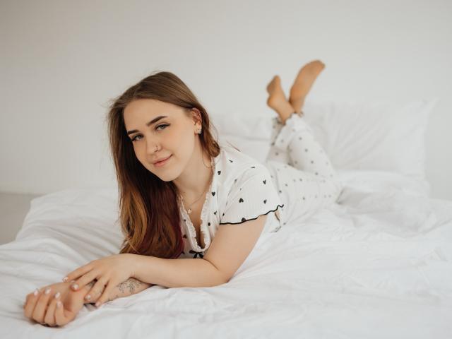 LauraHlot, eine attraktive Frau mit langen braunen Haaren, liegt auf einem weißen Bett, trägt ein weißes, gepunktetes Hemd und hat die Beine angezogen. Füße in der Luft, lächelnd, weiches Licht, entspannte Pose..
