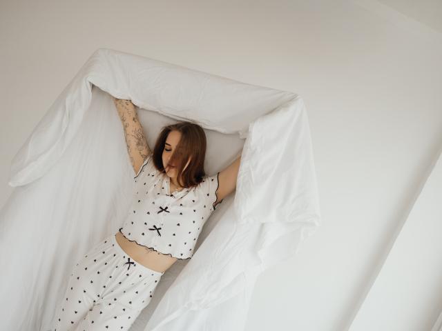 LauraHlot liegt in einem weißen Bett mit schwarzen Sternenmustern, die Arme sind erhoben, die tätowierten Unterarme sind sichtbar, ein weißes Laken ist teilweise heruntergezogen. ihren Körper bedeckend, weiches Licht, entspannte Pose, minimalistische Umgebung.