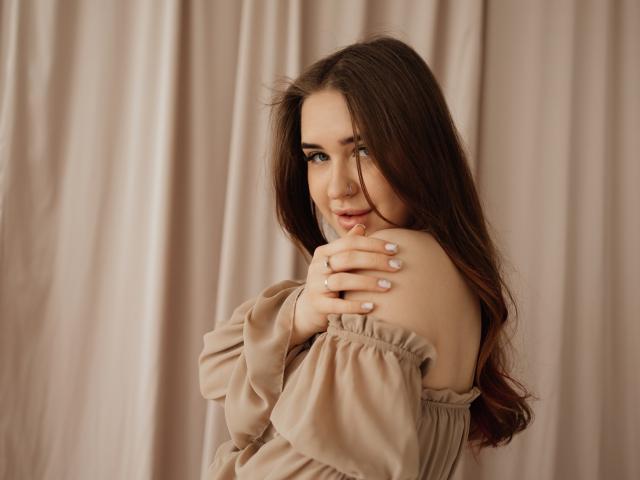 Photographie de LauraHlot, avec de longs cheveux bruns, regardant par-dessus son épaule, les mains sur son épaule, portant une haut beige à épaules dénudées, lumière douce, léger sourire, sexy, flou artistique, fond de rideau..