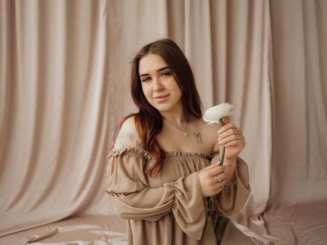 LauraHlot tiene un fiore di margherita bianco nella mano destra, e indossa un abito beige con spalline scoperte e dettagli in pizzo. Ha lunghi capelli castani, pelle chiara e un sorriso delicato. Incredibilmente sexy..