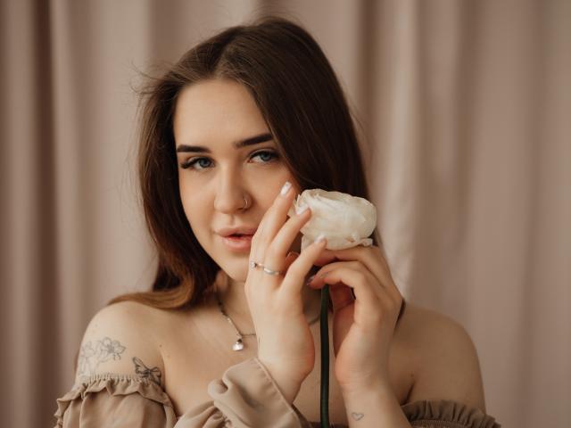LauraHlot mit dunkelbraunen Haaren, die einen mit weißem Zuckerguss überzogenen Donut nahe an ihr Gesicht hält, trägt ein beiges Oberteil mit einem Schulterauschnitt. sichtbares Tattoo an der rechten Schulter, Nasenring, verführerischer Blick, weiches Licht.