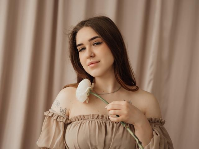 LauraHlot, fair-skinned, with long brown hair, holds a white rose to her chest. She wears an off-shoulder beige dress with lace detailing, looking slightly smug. Soft, muted background.