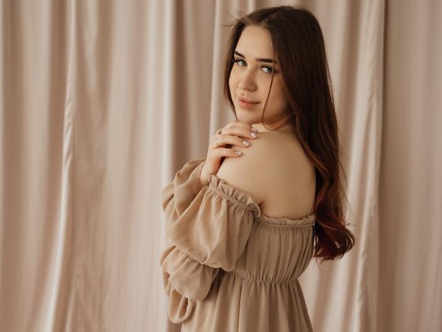 LauraHlot se tient devant des rideaux beiges, vêtue d'une robe beige à une épaule, avec des manches bouffantes. Elle a de longs cheveux bruns, des yeux bleus et un sourire timide qui lui effleure la clavicule. Extrêmement séduisant..